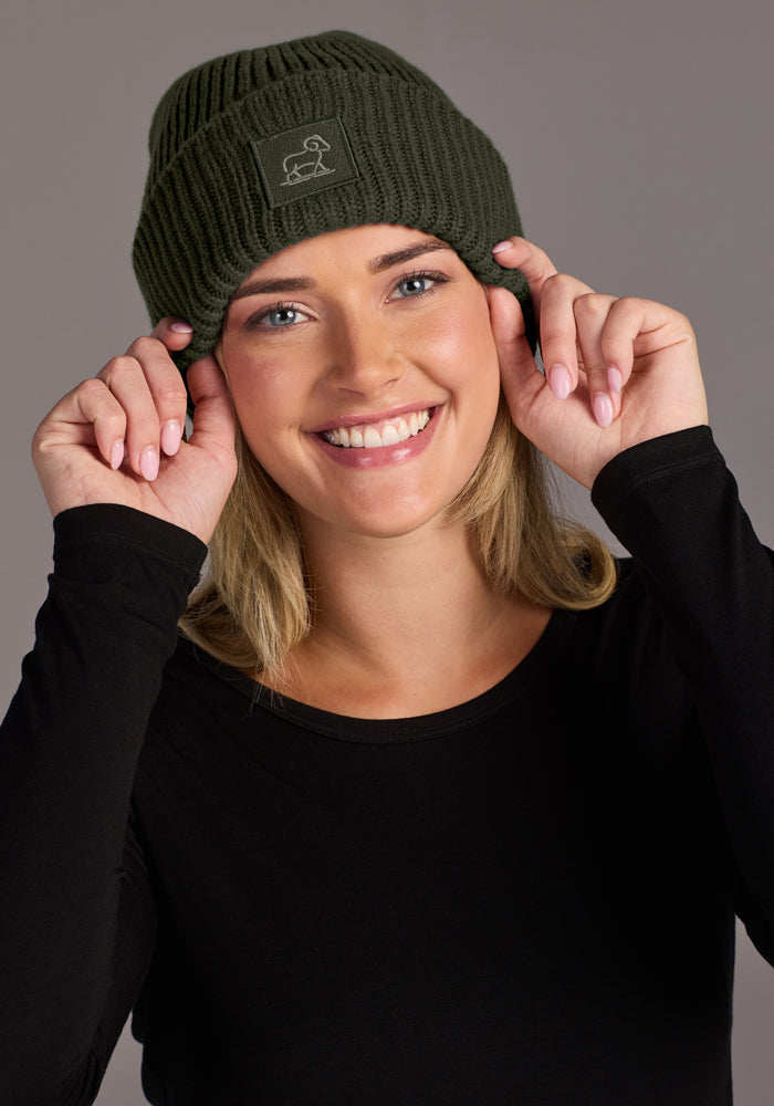 Smiling person in black top adjusts Woolx Ria Chunky Beanie - Duffel Bag, hands on hat, plain gray background.