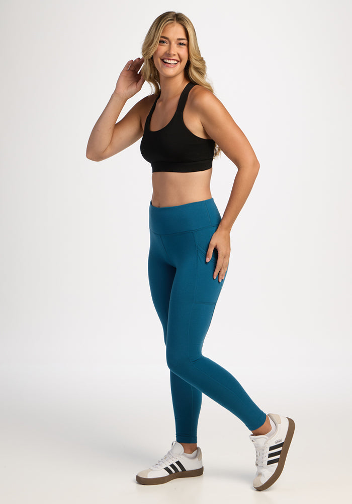 Smiling woman in Woolx Piper Pocket Leggings - Blue Pine, black sports bra, and sneakers poses on a white background.
