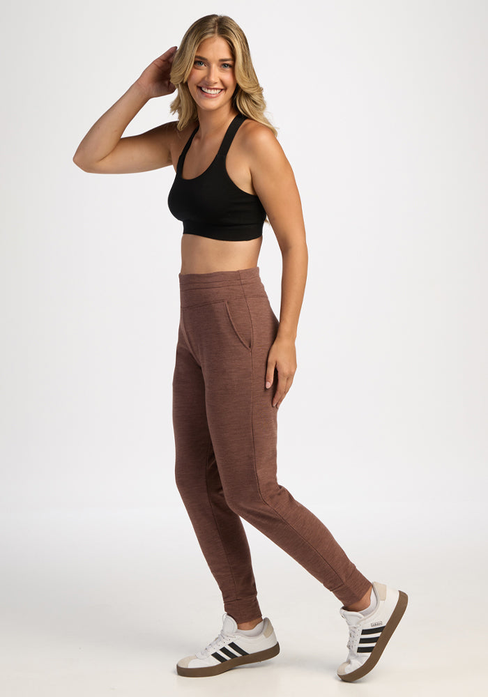 Smiling woman in Woolx Willa Joggers - Sable Heather, black sports bra, and white sneakers stands against a white background.