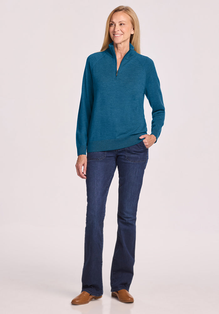 Woman smiles, hand in pocket, wearing Woolx Collins Half Zip Sweater - Blue Pine, blue jeans, and brown shoes.