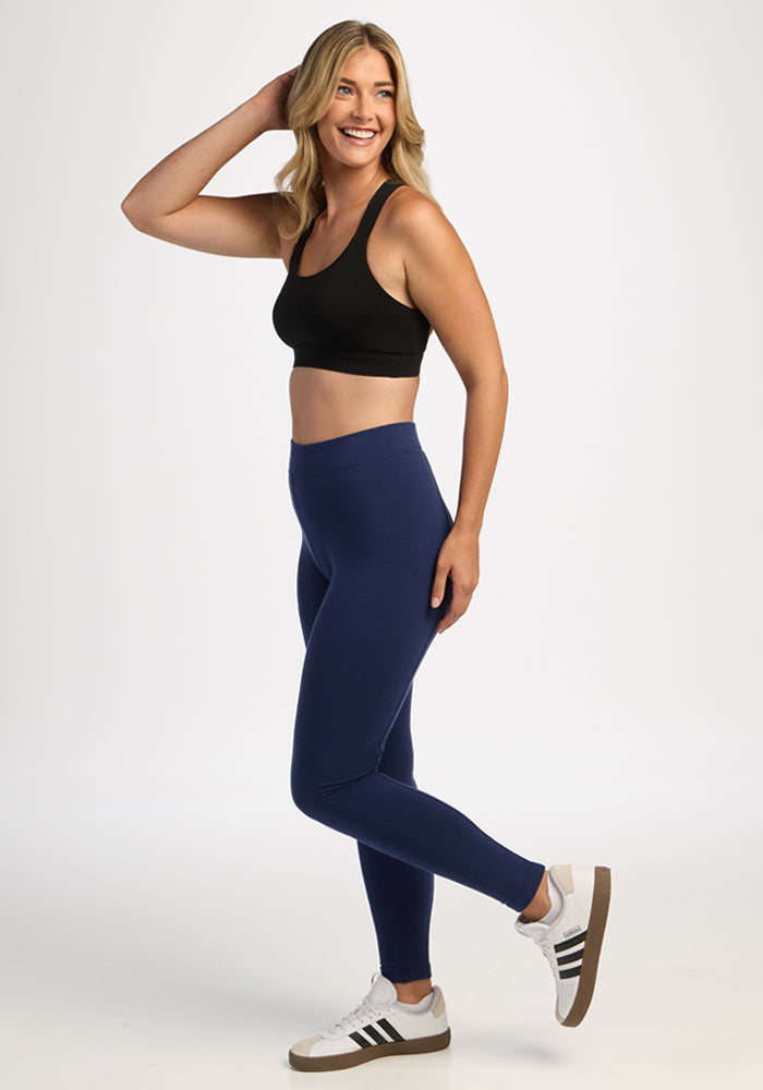 Smiling woman wears Woolx Stella Leggings - Ocean Cavern and a black sports bra, standing on one leg, plain backdrop.