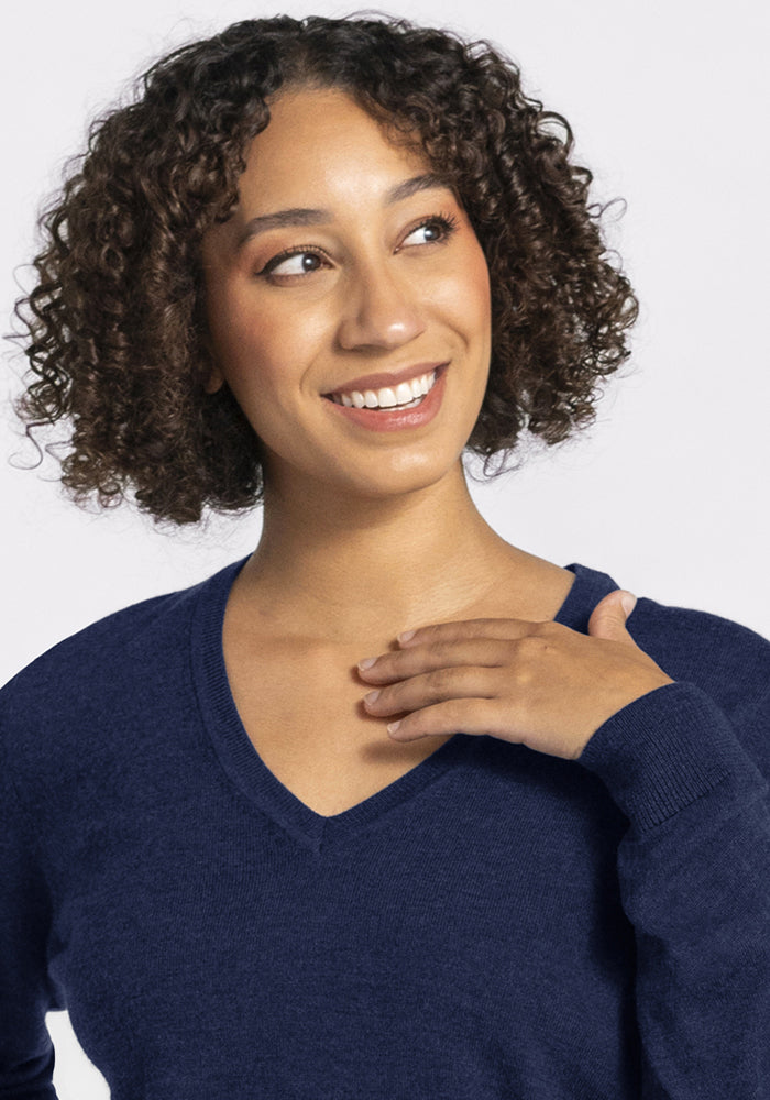 Smiling person with curly hair in Woolx Francesca Sweater - Ocean Cavern, hand to chest, looking to the side.