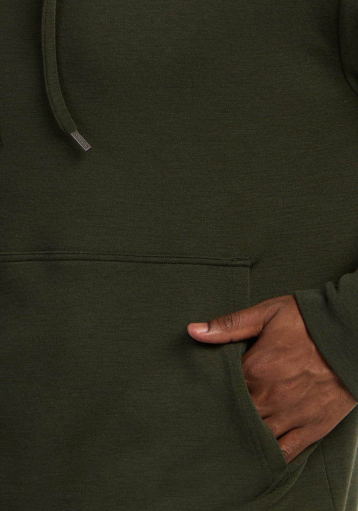 A hand rests in the front pocket of the Woolx Watson Pullover - Duffel Bag, an odor-resistant Merino hoodie.