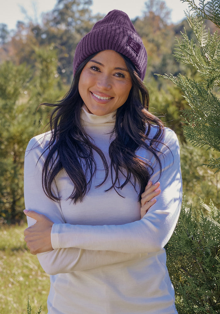 Woman in Woolx Ria Chunky Beanie - Purple Iris and white turtleneck smiles outdoors among sunlit green trees.