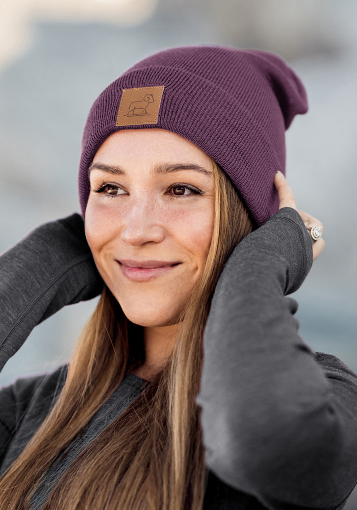 Woman smiles, wearing Woolx Baylor Beanie in Purple Iris and a gray sweater, holding her cozy winter hat.