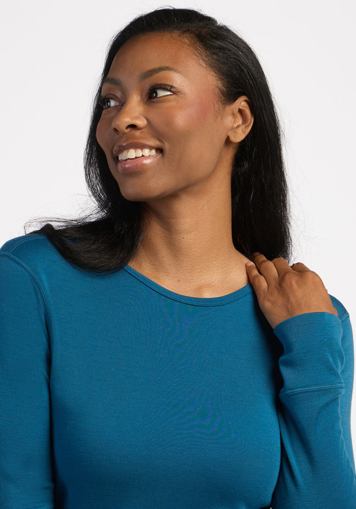 Woman in a Woolx Hannah Long Sleeve Top - Blue Pine smiles, looking left—a perfect moisture-wicking base layer.
