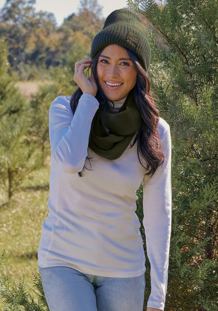 Woman in a green beanie and Woolx Noelle Infinity Scarf in Duffel Bag smiles outdoors among sunny green trees.