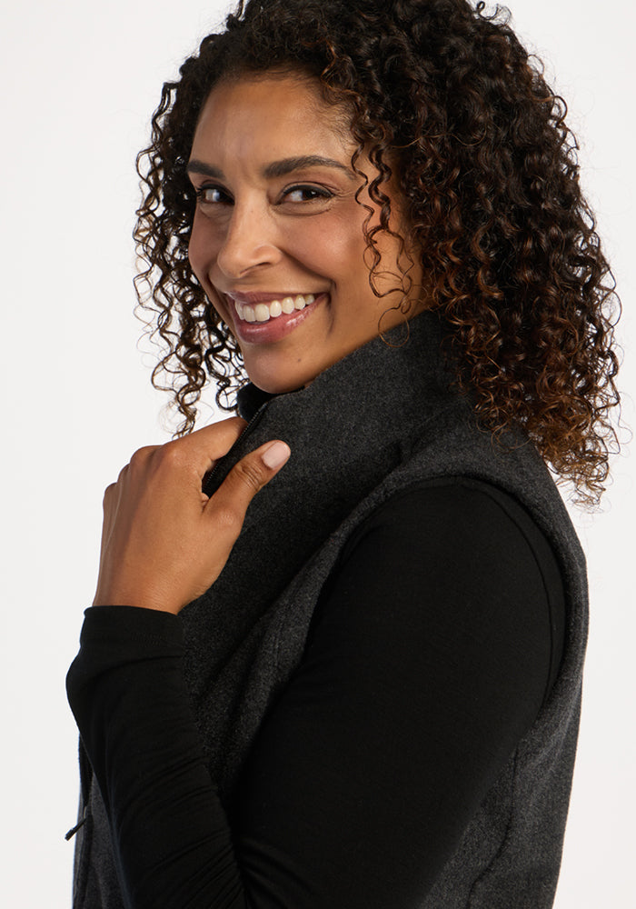 Woman with curly hair smiles over her shoulder, wearing the Woolx Haley Vest in Carbon Black against a light background.