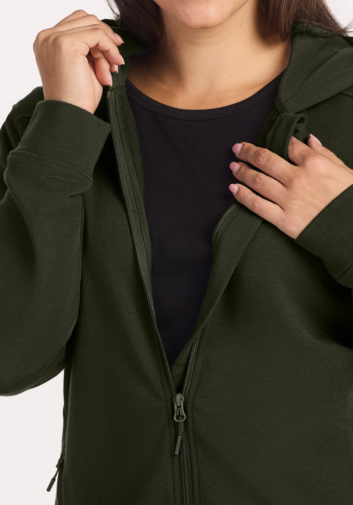Woman in a Woolx Darby Zip Up Hoodie - Duffel Bag, unzipping the cozy dark green hoodie with one hand at her collar.