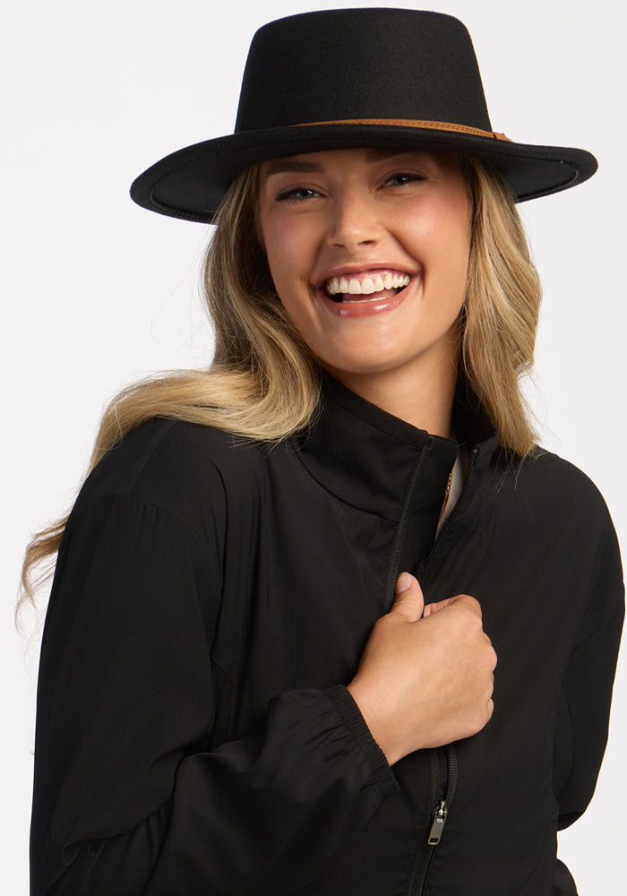 Smiling woman in Woolx Cooper Windbreaker - Black and hat, holding her collar against a white background.