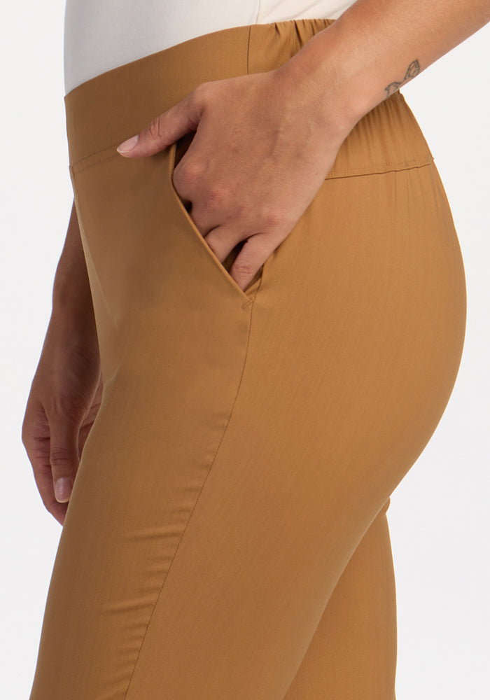 A person wears Woolx Nicole Tall Ankle Pant in Saddleback, a tan merino wool style, hand in pocket, waist to mid-thigh.