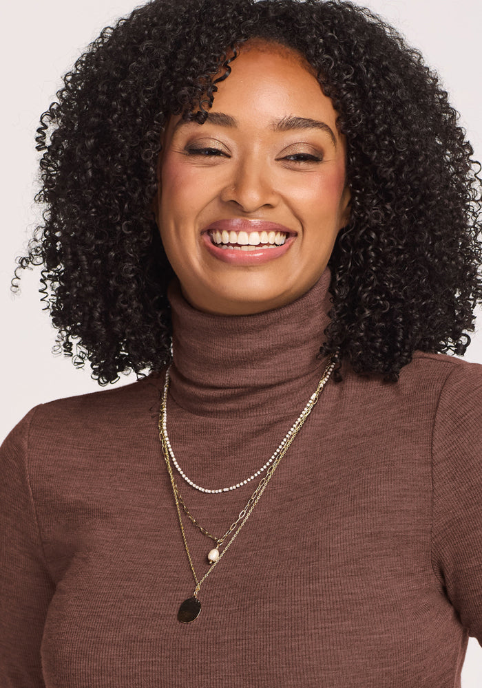 Smiling woman with curly hair wears Woolx Sage Ribbed Turtleneck in Sable Heather, layered necklaces.