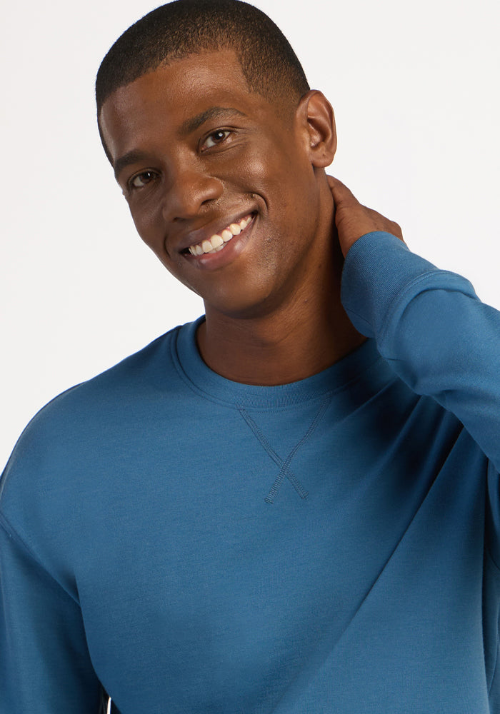 A smiling man in a blue ashes Miles Crewneck, a heavyweight, moisture-wicking merino wool sweater with a V-stitch collar.