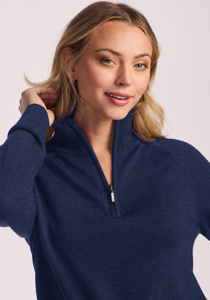 Woman with long blonde hair wears Woolx Collins Half Zip Sweater in Ocean Cavern, smiling and touching her hair.
