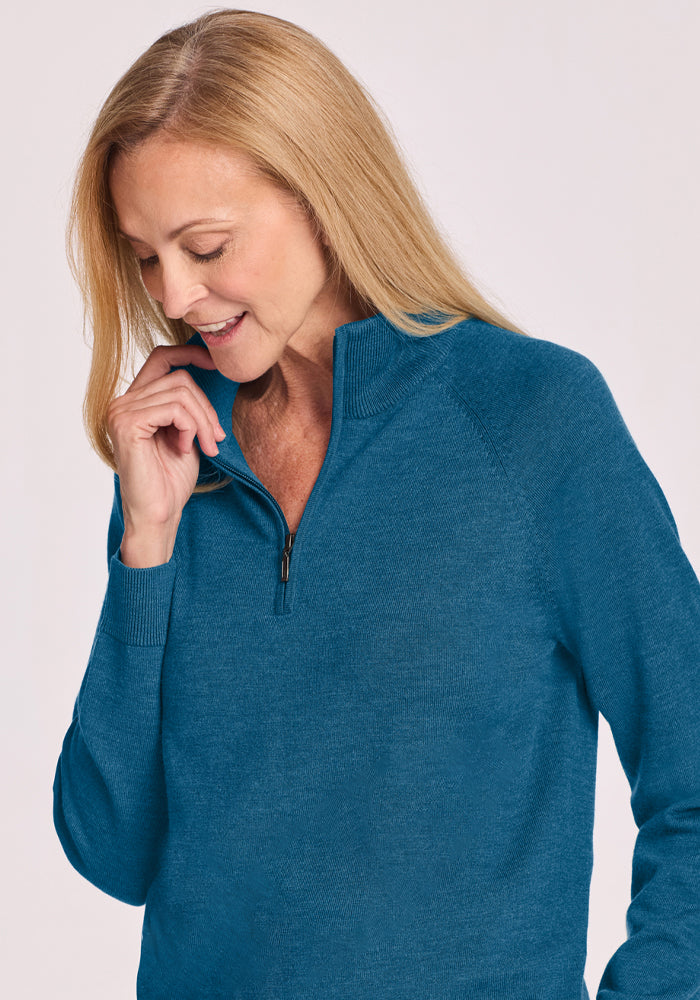 Blonde woman in Woolx Collins Half Zip Sweater - Blue Pine looks down, touching the collar near her chin.