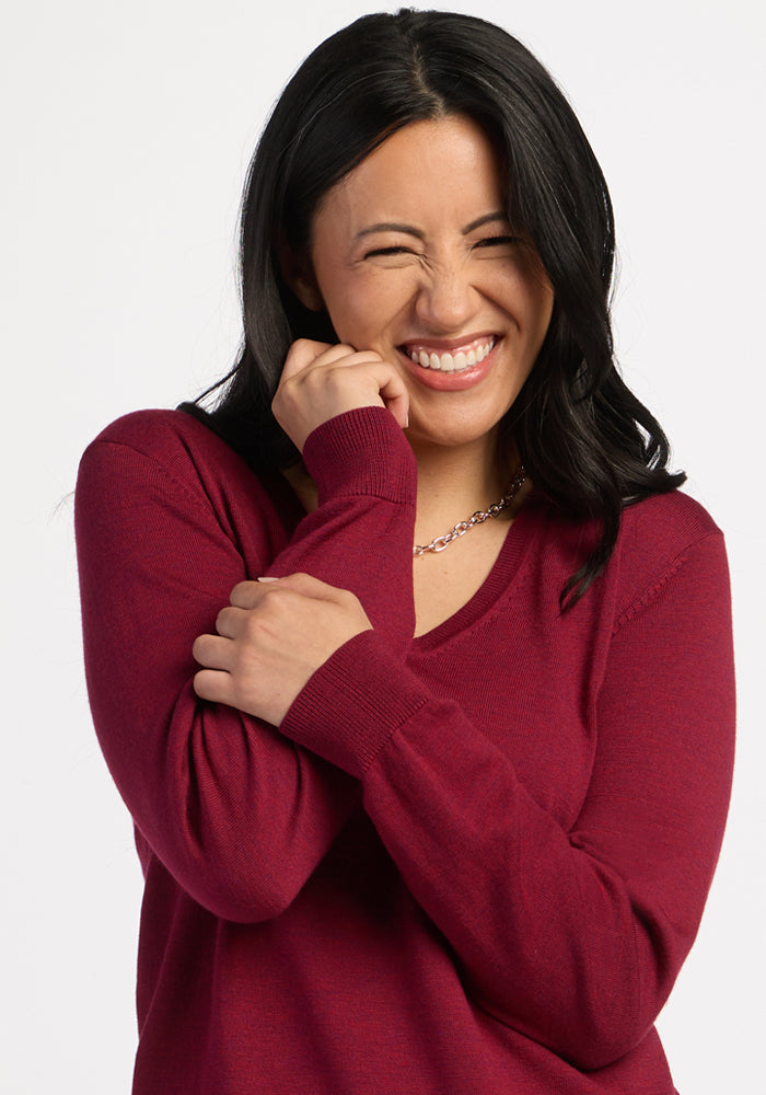 Smiling with eyes closed, she hugs herself in the Beet Red Francesca Sweater by Woolx—happy and travel-ready.