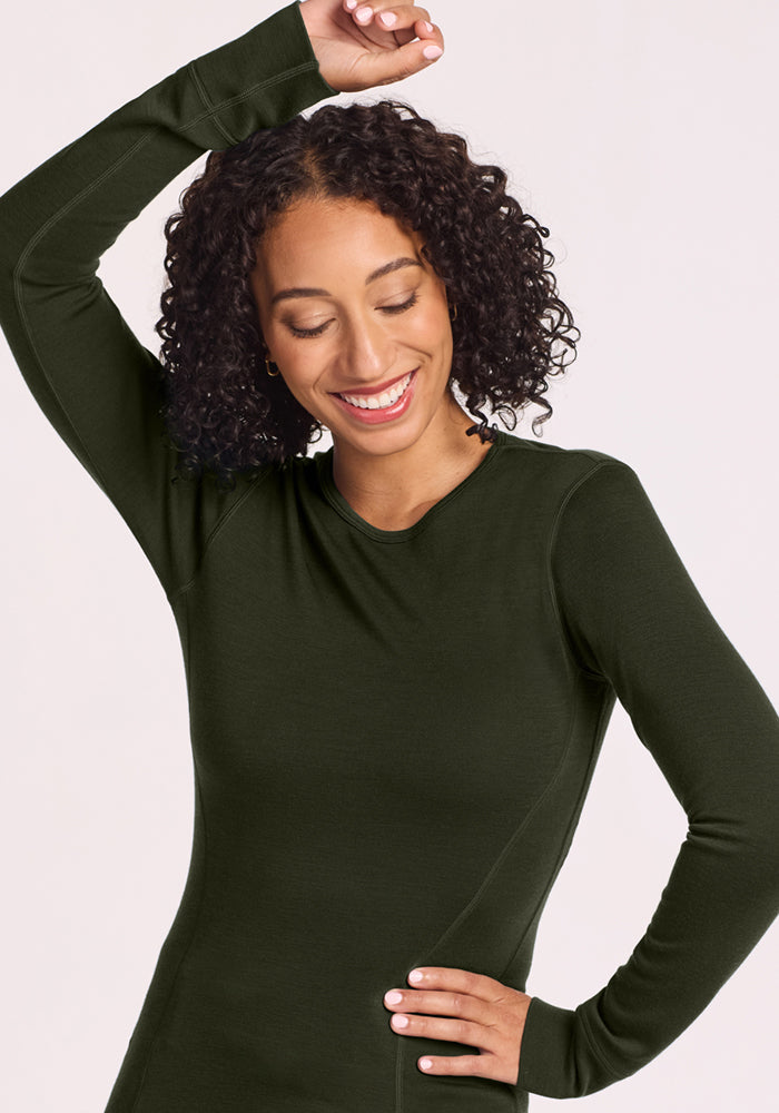 Smiling woman with curly hair wears Woolx Hannah Long Sleeve Top in Duffel Bag, arm raised, on a light background.