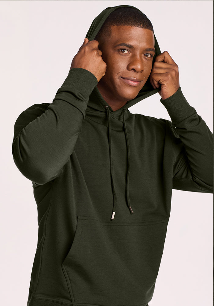 Man in a Woolx Chase Hoodie - Duffel Bag smiles, holding the hood with both hands against a plain light background.