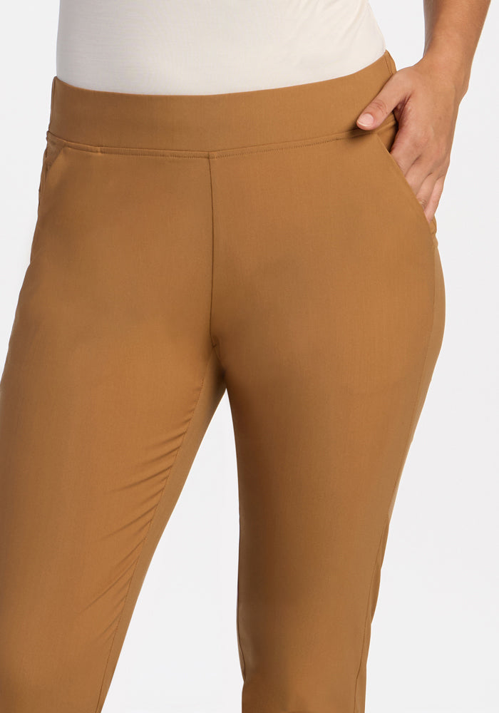 Model wears Woolx Nicole Tall Ankle Pant in Saddleback tan with a white top, one hand in pocket, on plain background.