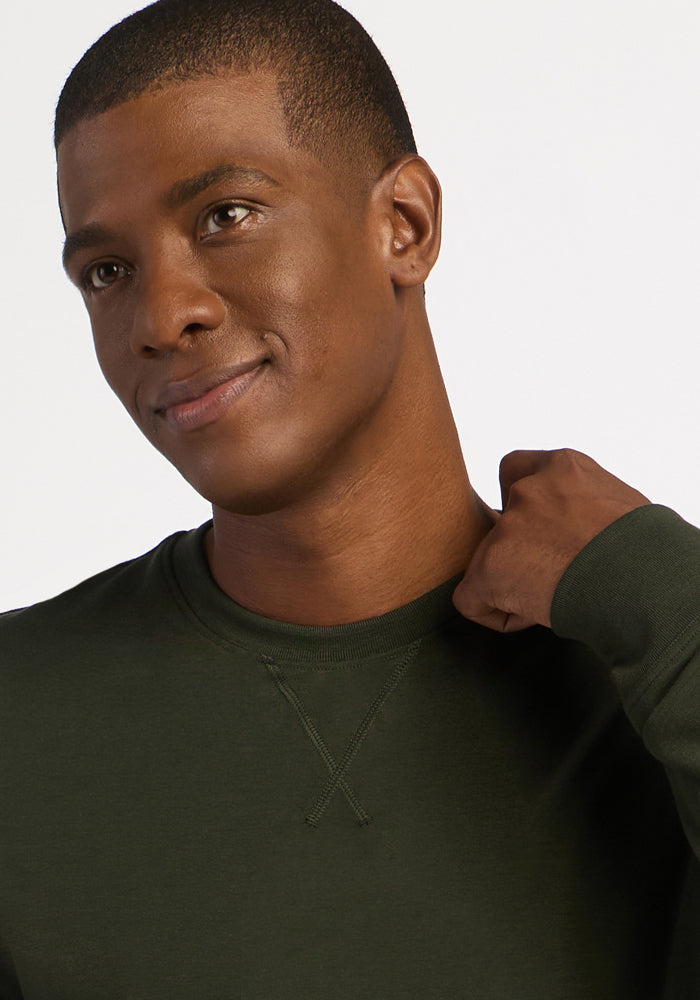 A man wears a dark green Woolx Miles Crewneck - Duffel Bag, a heavyweight merino sweater with a V-stitch collar detail.