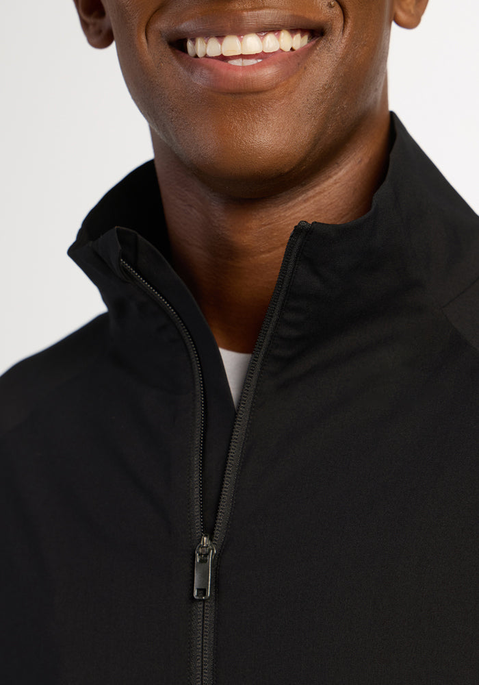 A person smiles, shown from the shoulders up, wearing a Woolx Dominator Windbreaker in black with the collar partially up.