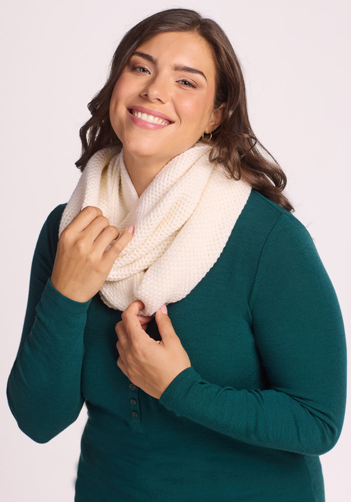 A smiling woman with long brown hair wears a green top and the Woolx Noelle Infinity Scarf in Buttercream, holding the cozy accessory with one hand against a plain light background.