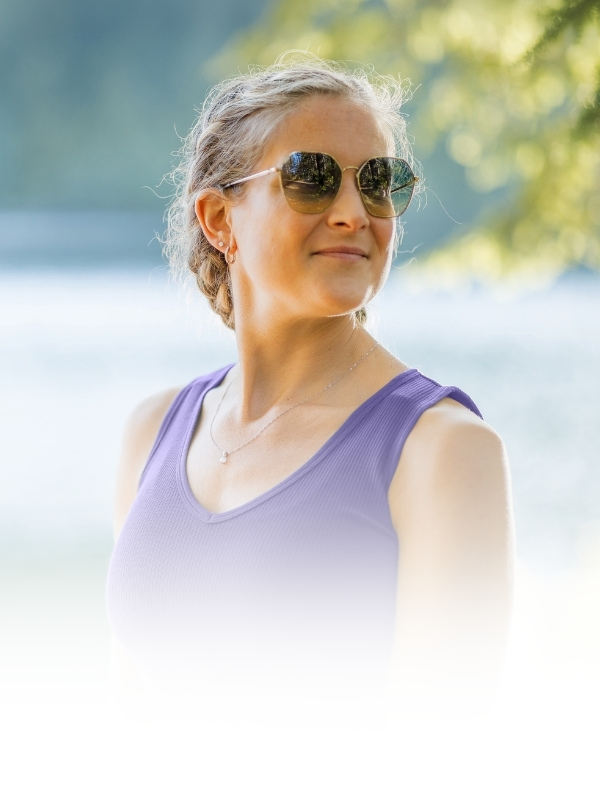 A woman outdoors wearing round dark sunglasses and a lavender-purple ribbed tank top, with a soft blurred natural background.