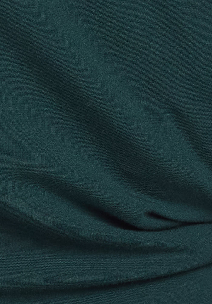 A close-up of Woolx Stella Leggings in Deep Timber shows dark green fabric with visible weave texture and gentle folds, reminiscent of cozy merino wool, creating soft shadows and highlights.