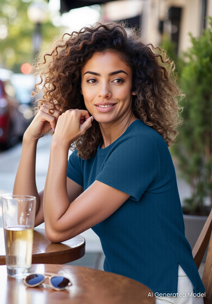 A woman with curly hair wears the Woolx Delia Ribbed Short Sleeve in Moroccan Blue as she sits smiling at an outdoor café table, with a glass of water and sunglasses beside her. Trees and a blurred street are visible in the background.