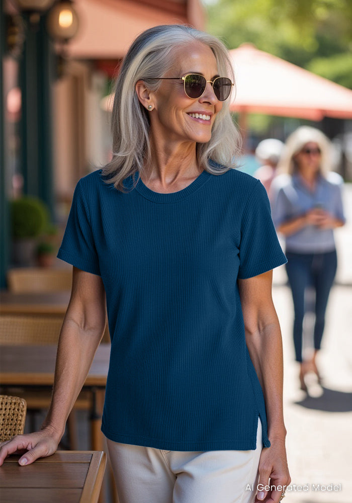 A smiling older woman with gray hair and sunglasses wears the Woolx Delia Ribbed Short Sleeve in Moroccan Blue and light pants, standing by an outdoor café table on a sunny day; another person walks in the blurred background.