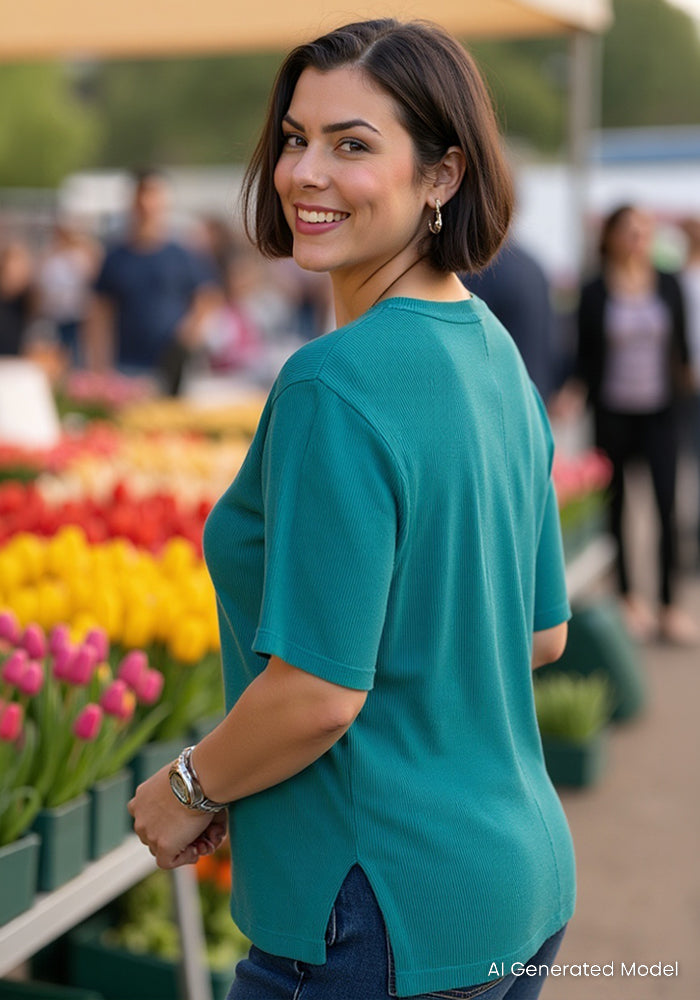 A woman with short dark hair, wearing the Woolx Delia Ribbed Short Sleeve in Surfside, smiles in front of colorful tulip bouquets at an outdoor market with blurred people and booths behind her.