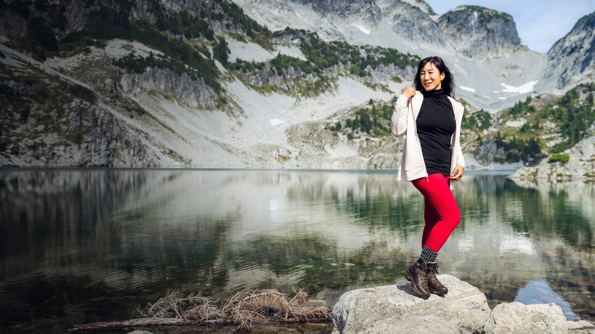 A woman standing on the shore of a mountain side lake wearing bright red leggings and a zip up hoodie.