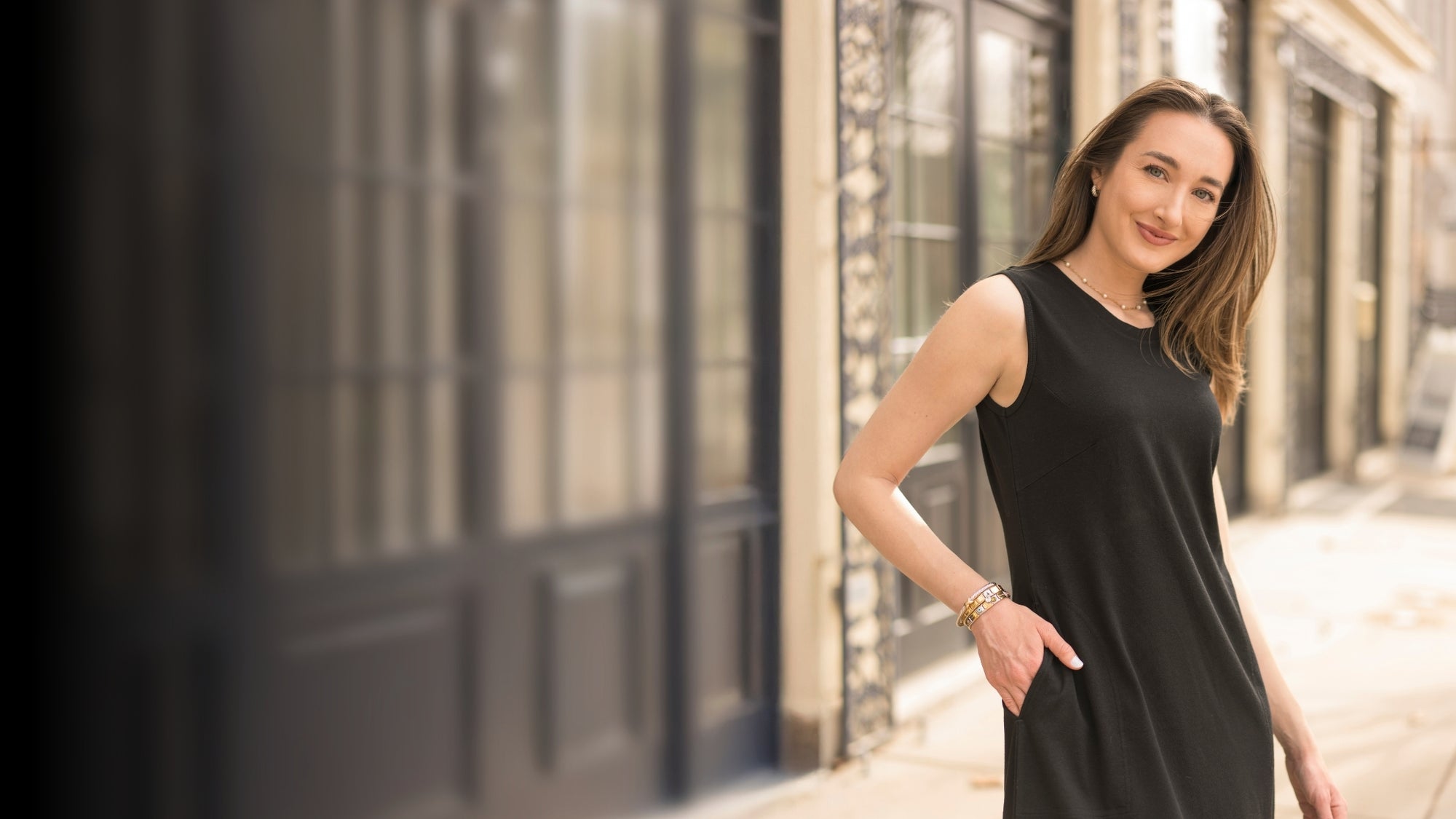 A woman wearing a sleeveless black dress stands on a city sidewalk in front of a blurred building facade with dark windows.