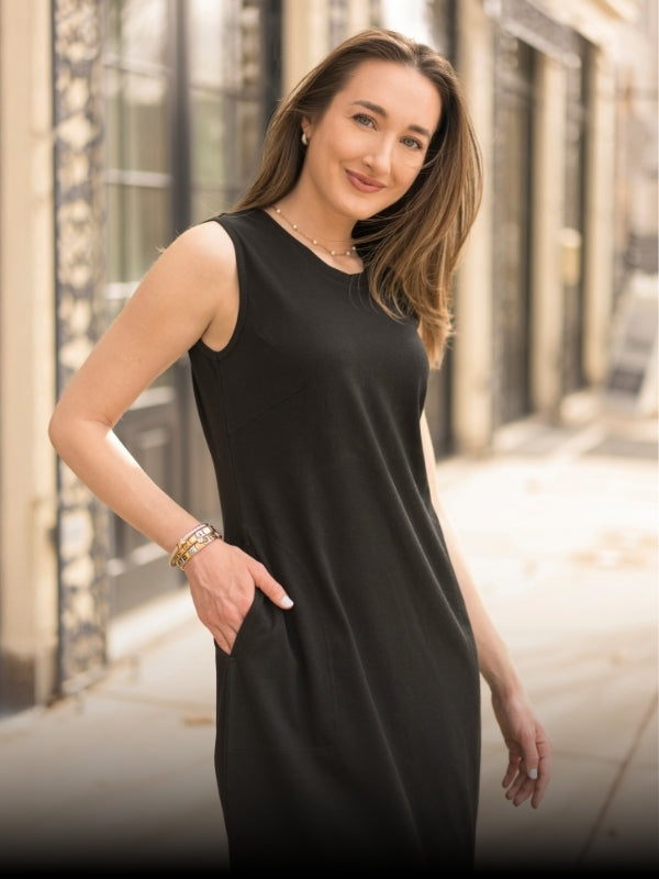 A woman wearing a sleeveless black dress with side pockets, standing outdoors on a city sidewalk with blurred buildings in the background.