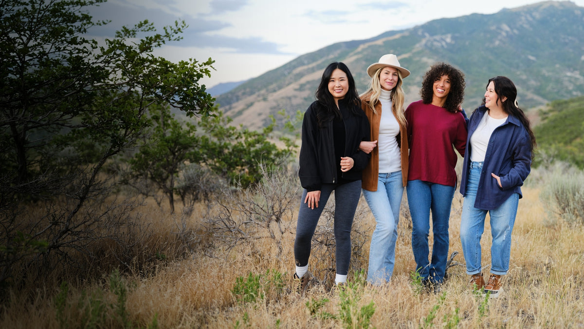 Four women wearing colorful wool layers pose together in a grassy field with mountains in the background.