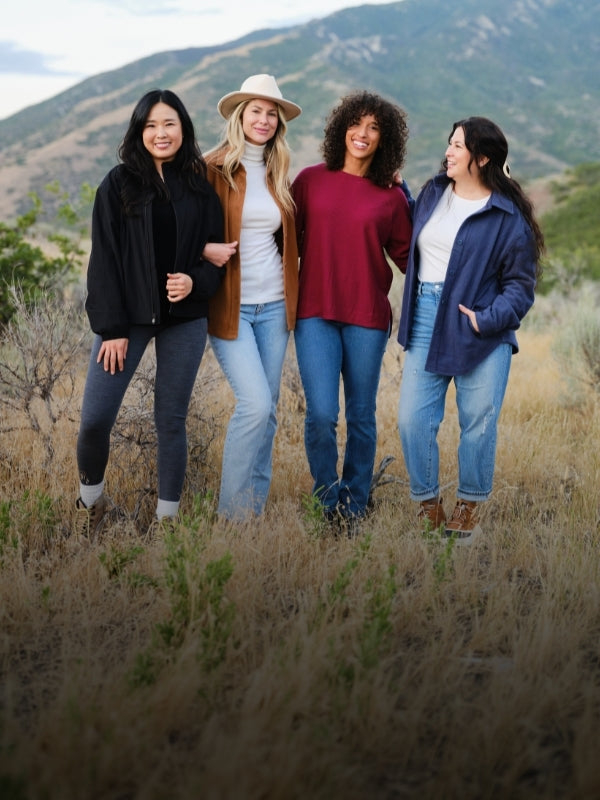 Four women wearing colorful wool layers pose together in a grassy field with mountains in the background.