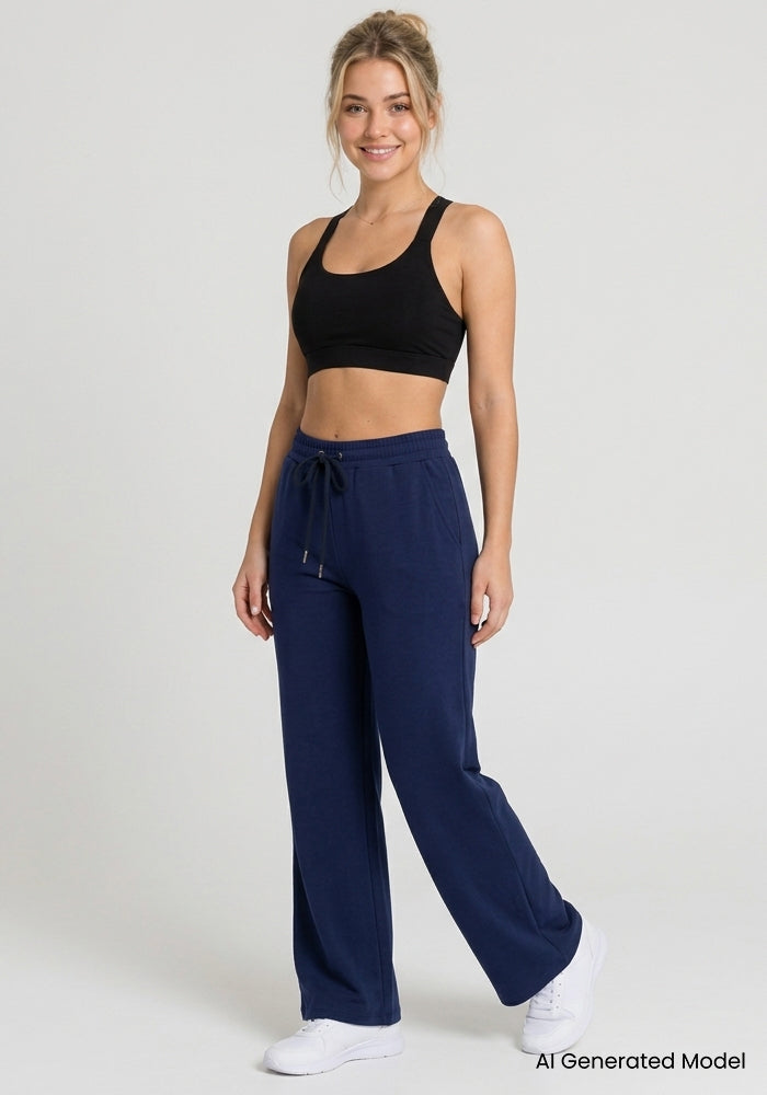 A woman in Woolx Eloise Petite Pants - Ocean Cavern smiles in a black sports bra and white sneakers against a plain backdrop.