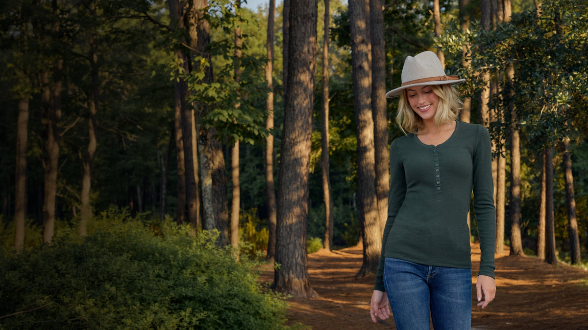 A woman wearing a dark green long-sleeve henley shirt, blue jeans, and a beige wide-brim hat stands on a forest path.
