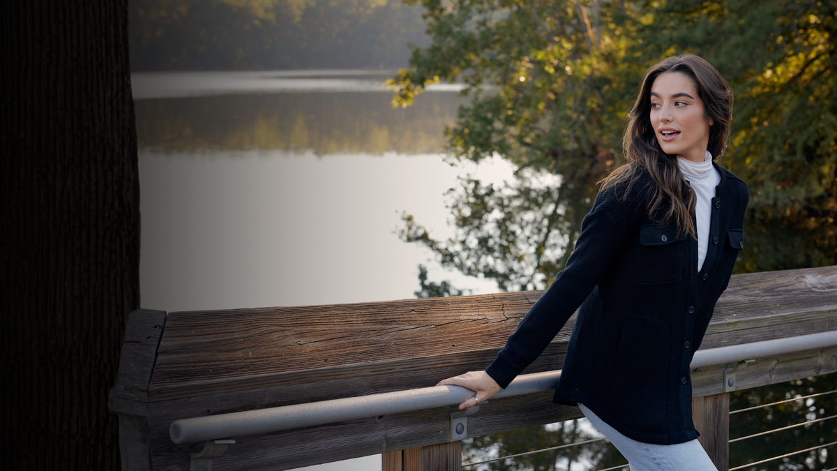 A woman wearing a black blazer over a white turtleneck and white pants leans against a wooden railing outdoors with a lake and trees in the background.