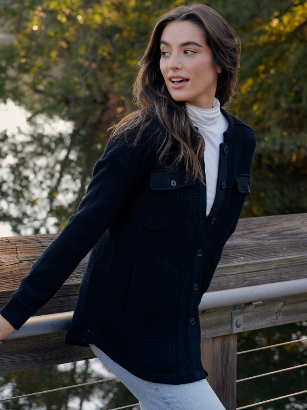 A woman wearing a black blazer over a white turtleneck and white pants leans against a wooden railing outdoors with a lake and trees in the background.
