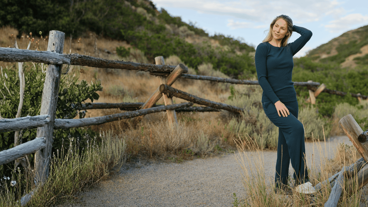 A woman wearing a dark teal long-sleeve top and matching wide-leg pants stands on a dirt path outdoors near a rustic wooden fence and hills.