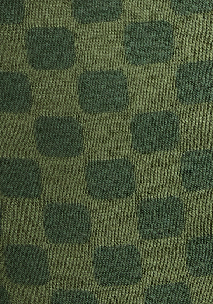 Close-up of the Woolx Kids Explorer Base Layer Top - Frog Hop, showing its breathable Merino wool fabric in a green and dark green checkerboard pattern with oval and rectangular shapes. The knit texture is clearly visible.