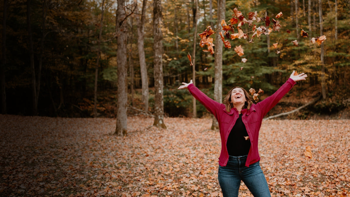 A smiling woman in a magenta cardigan tosses autumn leaves into the air in a wooded clearing.