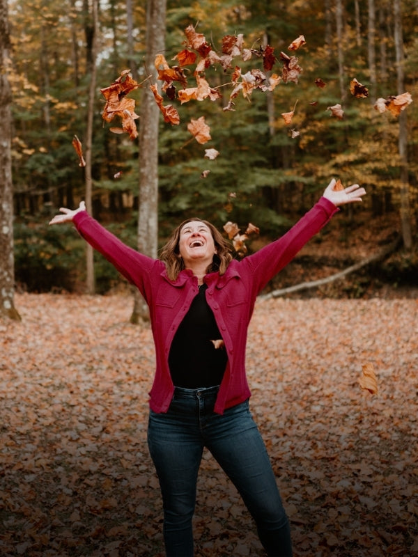 A smiling woman in a magenta cardigan tosses autumn leaves into the air in a wooded clearing.