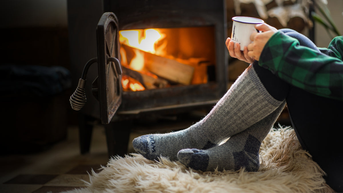 Person sitting in front of a small stove wearing Extreme socks and holding a mug