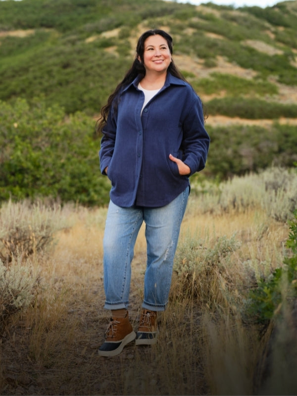 A woman standing in a field wearing a blue shacket, blue jeans and boots. 