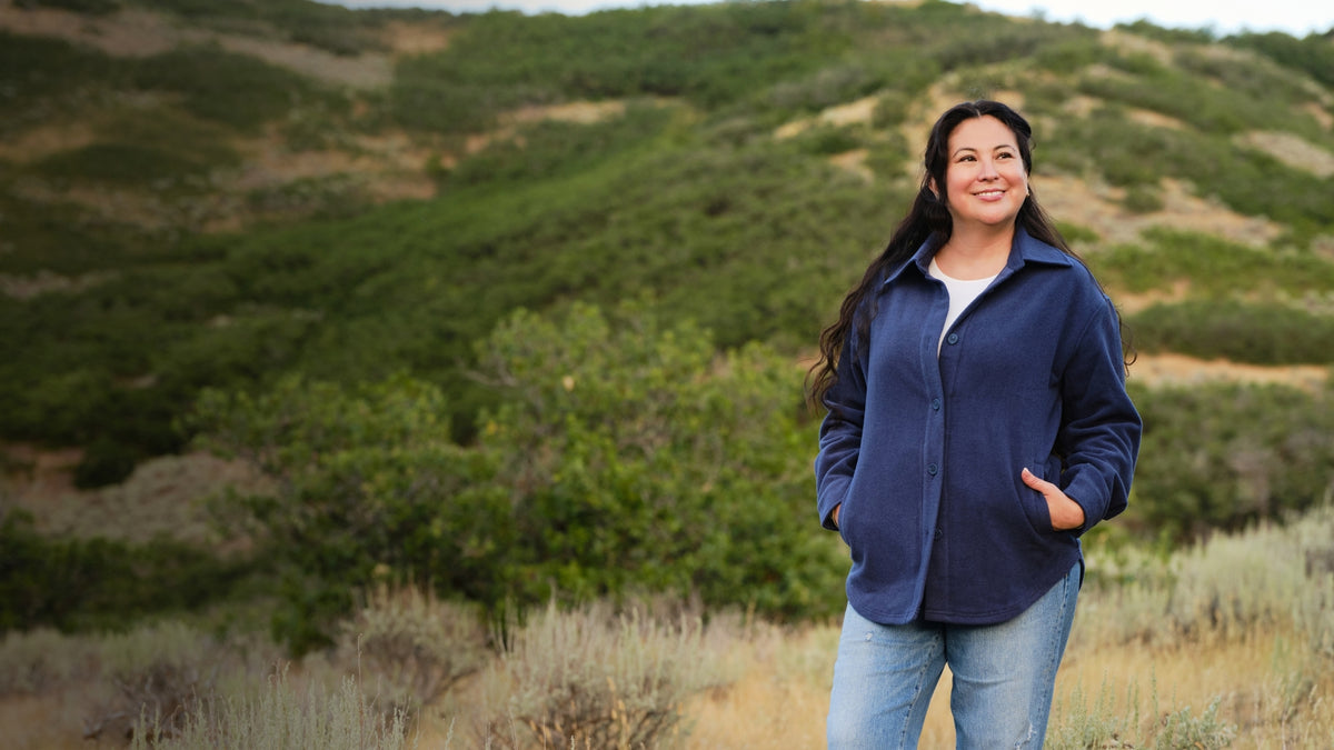 A woman standing in a field wearing a blue shacket, blue jeans and boots. 