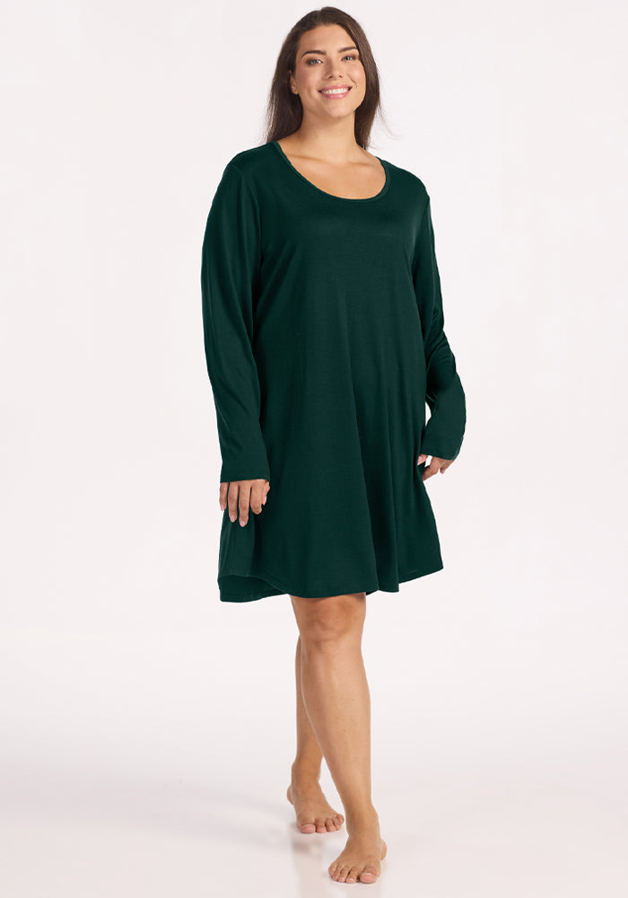 A smiling woman wears a dark green Lucille Long Sleeve Nightgown - Deep Pine that hits above the knee.