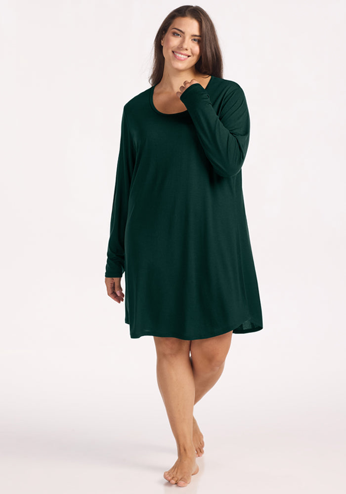 A smiling woman wearing a dark green long-sleeve Woolx Lucille Long Sleeve Nightgown - Deep Pine. 