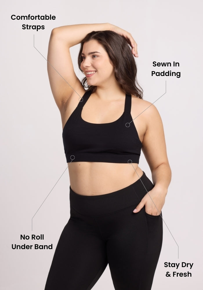 A smiling woman wears the Woolx Livi Bra - Skyline in black with leggings, raising one arm. Labels highlight its comfy straps, sewn-in padding, no-roll under band, and stay-dry & fresh material.